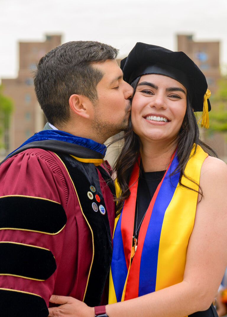 Man kissing a graduate on the cheek, she wears a cap, gown and yellow stole