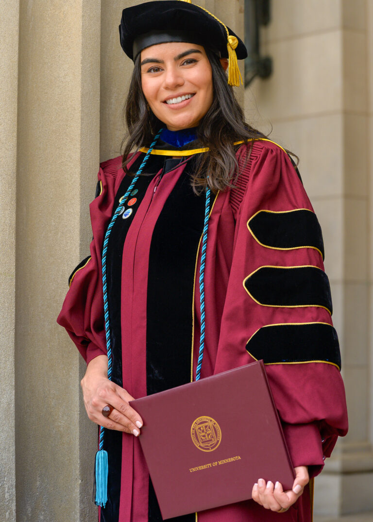 Graduate in a cap and maroon doctoral gown holding a diploma cover in front of a stone column