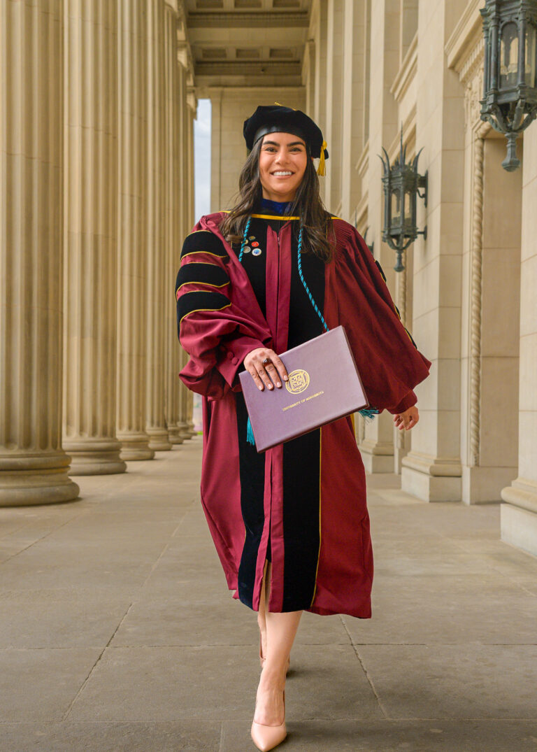 Graduate in a cap and maroon gown walking between stone columns holding a diploma