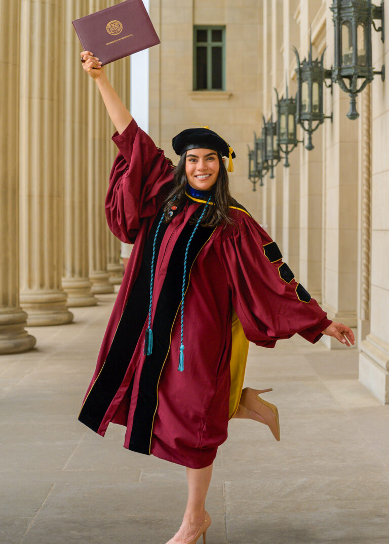 Graduate in a cap and maroon gown celebrating with a diploma raised beside stone columns