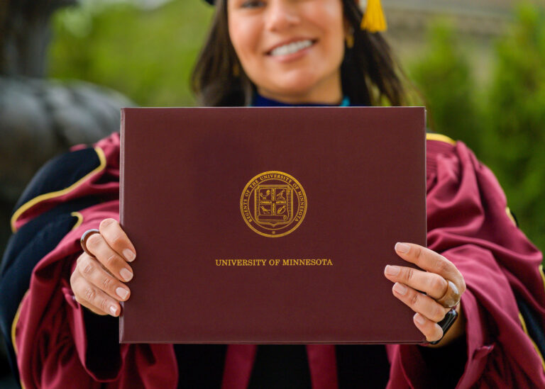 Graduate in academic regalia smiling while holding a maroon diploma cover