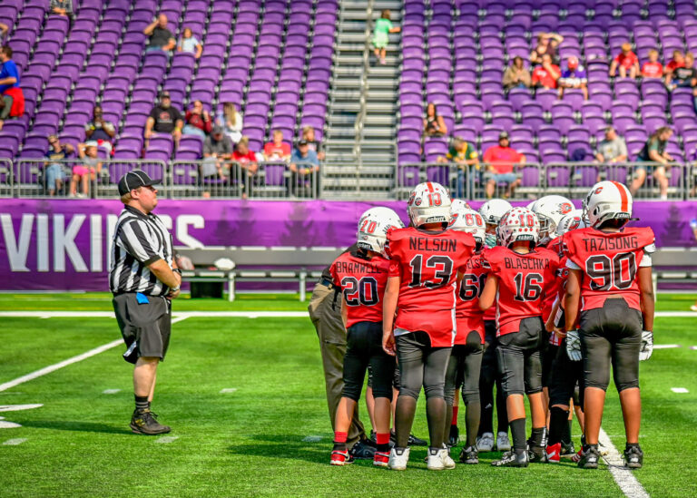 Youth football team in red uniforms huddled on the field with a referee nearby
