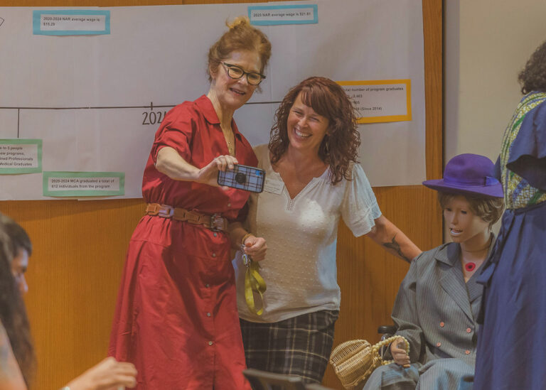 Two women smiling for a selfie in front of a timeline poster at an anniversary event