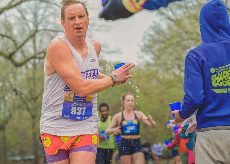 Runner in a white tank top taking a cup of water along the course with other runners behind