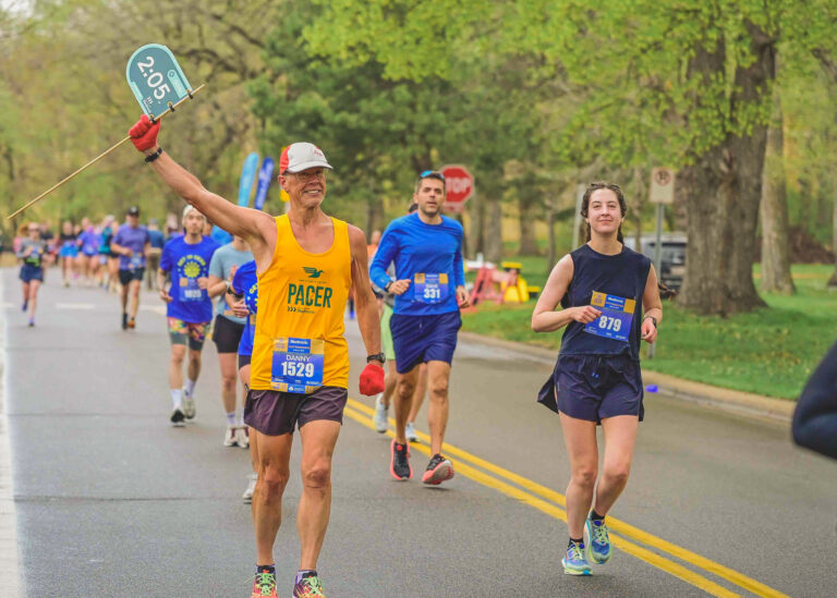 Runner in a yellow shirt raising an arm along a tree-lined race course