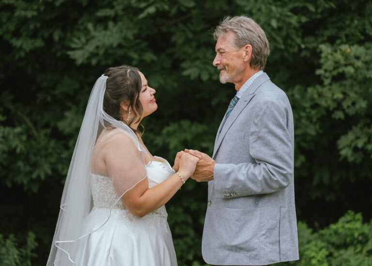 Bride in a veil holding hands with an older man in a gray suit on a lawn
