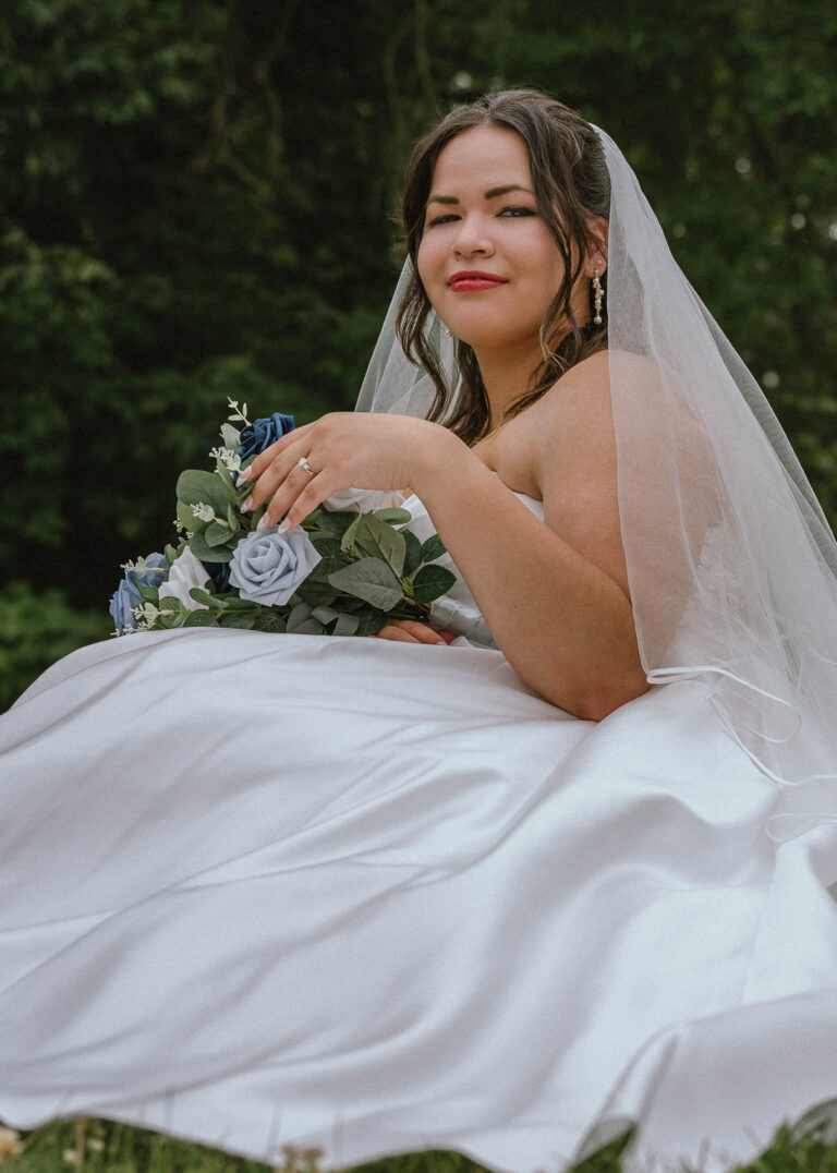 Bride in a white dress and veil sitting on the grass holding a blue and white bouquet