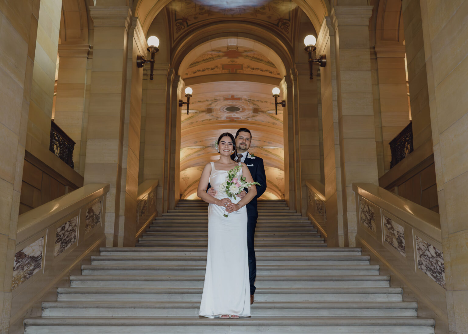 Bride and groom standing together at the foot of a grand marble staircase