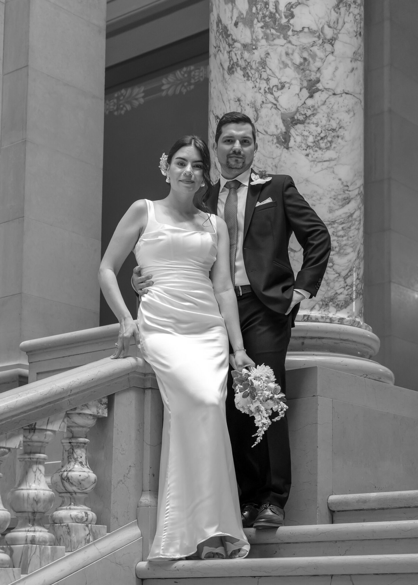 Bride and groom leaning on a stone balustrade beside a marble column, in black and white