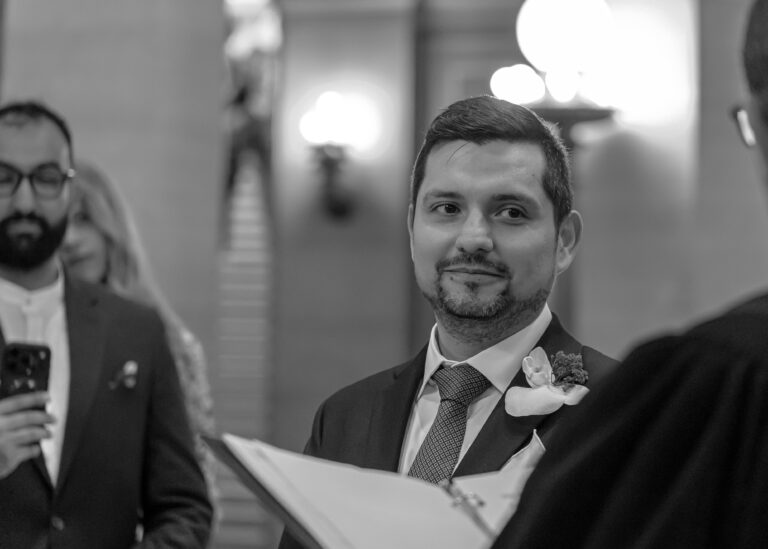 Groom smiling during the ceremony with guests behind him, in black and white